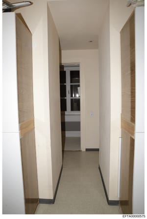 This is a photograph documenting the interior of a hallway within a property, showing clean, modern finishes with cream-colored walls, light wood doors on either side, and what appears to be a glass-paneled door or window at the end of the corridor. 