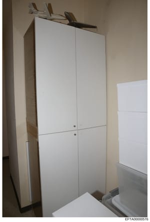 This is an evidence photograph showing a white four-door cabinet or wardrobe unit installed in what appears to be a residential interior space. The cabinet features white laminate doors with metal handles and appears to be positioned against beige/cr