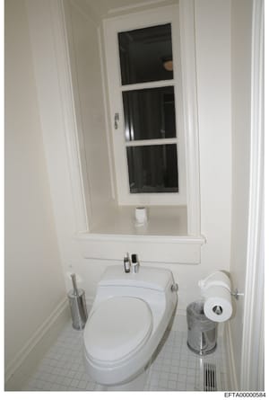 This is a photograph of a residential bathroom interior, showing a white toilet, pedestal sink, medicine cabinet with mirror, and basic bathroom accessories. The image appears to be documentation of a property's interior, likely taken as part of the 