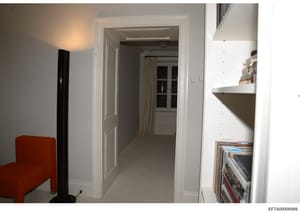This is an evidence photograph showing an interior hallway or corridor space, likely from one of Jeffrey Epstein's properties. The image depicts a modern residential interior with white walls, white trim, a red upholstered chair, built-in shelving, a
