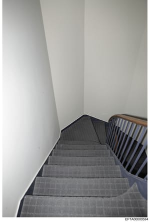 This photograph shows an interior staircase viewed from above, featuring gray carpeted steps and a wooden handrail with blue-striped spindles. The stairs appear to be in a residential or commercial property. The image is taken from a downward angle, 