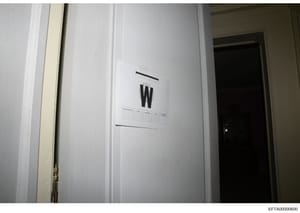 This is an evidence photograph from a property search or investigation showing an interior wall with an evidence marker labeled 'W' and what appears to be a small evidence documentation label or grid below it. The marking and documentation protocol s