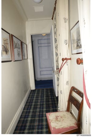 This is a photograph of a narrow residential hallway featuring cream-colored walls, a blue door at the far end, a plaid/tartan pattern floor runner, framed artwork on the walls, and decorative elements including wooden chair details visible on the ri