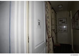 This is an evidence photograph showing an interior hallway of a property, featuring a white door marked with an 'X' and what appears to be evidence documentation. The image captures the hallway's architectural details including decorative wallpaper, 