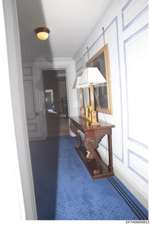 This photograph documents an interior hallway space, showing a decorated wooden console table with a framed mirror, table lamp, and decorative items. The hallway features white paneled walls, a blue patterned carpet, and recessed ceiling lighting. Th