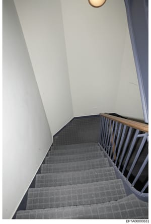 This is a photograph of an interior stairwell featuring gray carpeted stairs with a wooden and metal railing. The image shows clean, institutional-style finishes with white walls and minimal decoration. The photo appears to be part of property docume