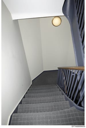 This is a photograph of an interior stairwell, showing a modern residential space with white walls, gray carpeted stairs, a wooden handrail, and brass wall sconce lighting. The image appears to be evidence documentation of property interior, likely f