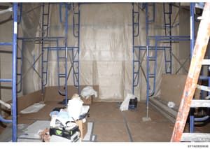 This photograph shows an interior space under construction or renovation, featuring blue metal scaffolding, plastic sheeting for containment, and construction materials scattered throughout. The image appears to be evidence documentation from a prope