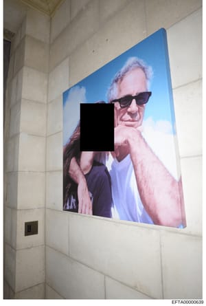 This is an evidence photograph showing a large portrait canvas or print mounted on a cream-colored tiled wall in what appears to be an interior hallway or entryway. The portrait depicts a man wearing sunglasses with his face redacted by a black recta
