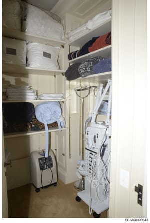 Evidence photograph showing the interior of a storage closet or linen room, containing shelves with bedding, linens, towels, and various household items. The photograph also documents white medical or electrical equipment stored on the right side of 
