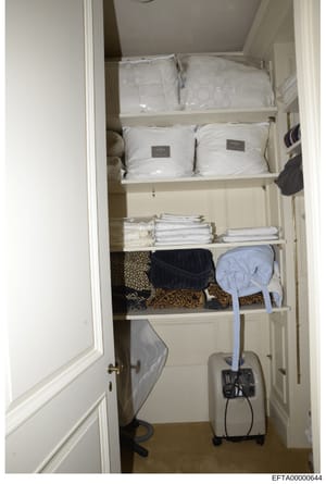 This is an evidence photograph of a residential linen closet, showing neatly organized shelving containing white bedding sets (some in plastic wrapping with labels), folded linens, decorative items, and various household goods. The photo appears to b