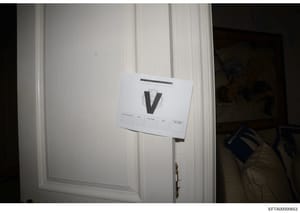 This is a forensic evidence photograph showing a white paneled door with a large black letter 'V' marker and an evidence documentation label attached. The marker and label system indicate this photo was taken as part of a systematic evidence collecti