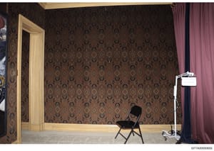 A photograph of an interior room showing ornate damask-patterned wallpaper in brown and gold tones, light wood trim, burgundy curtains, and minimal furnishings including a black folding chair and lighting equipment. The room appears to be photographe