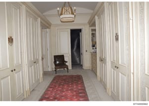 This is a photograph of an elegant hallway in a luxury property, featuring cream-colored paneled doors, a brass chandelier, red patterned runner rug, and classical furnishings. The image appears to be documentation of interior spaces within a propert