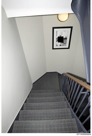This is an evidence photograph showing an interior stairwell with clean, minimalist décor. The image depicts gray carpeted stairs with a wooden handrail, white walls, a round brass wall sconce, and a framed black-and-white photograph on the wall (par