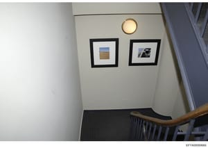 This photograph shows an interior wall space with decorative elements including two black-framed pictures hung on a light-colored wall, a round brass or gold-toned wall sconce light fixture positioned above the frames, and a wooden stair railing visi