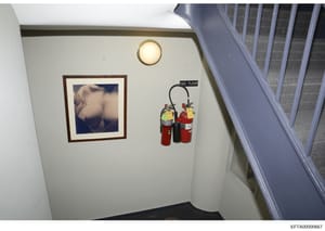 This is an evidence photograph showing the interior of a stairwell, likely from one of Jeffrey Epstein's properties. The image captures a cream-colored wall with a framed photograph, a circular ceiling light fixture, fire safety equipment, and a stai