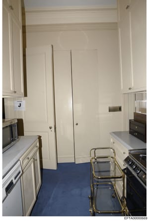 This is an interior photograph of a kitchen space showing white cabinetry, stainless steel appliances, and a blue floor. The image appears to be evidence documentation from a property investigation, showing the kitchen layout and condition. A marking