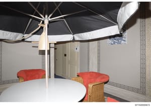 This is an evidence photograph showing an interior room with decorative furnishings, including a white round table, red cushioned chairs with wicker frames, and a large black and white umbrella. The room features patterned wallpaper with geometric de
