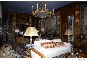 This photograph depicts an opulently decorated bedroom featuring high-end furnishings, including a crystal chandelier, wood paneling, fine art, and luxury bedding. The room appears to be part of a mansion or elite property, consistent with residences