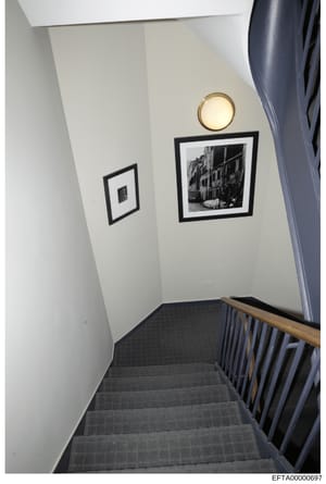 This is a photograph of an interior stairwell, showing a narrow landing or hallway area with cream-colored walls, dark carpeted stairs, and a wooden handrail. Two black-framed black-and-white photographs are displayed on the wall, along with a round 