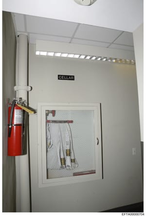 This is an evidence photograph showing the interior of a cellar room, featuring a red fire extinguisher mounted on the wall and electrical/mechanical equipment visible through a framed panel. The room is labeled 'CELLAR' and appears to be part of a p