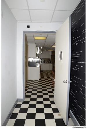 This is a photograph of a narrow hallway or corridor leading into a commercial kitchen or break room space. The image shows a black-and-white checkered tile floor, white walls with drop ceiling, and a doorway opening into a kitchen area with visible 