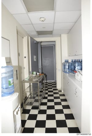 This photograph depicts an institutional-style hallway or break room with black and white checkered flooring, white drop-ceiling tiles, and commercial fixtures. The space contains water coolers, a stainless steel sink/counter unit, storage shelves wi
