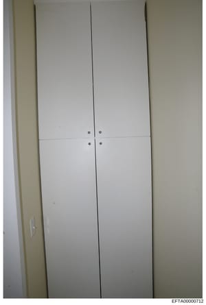 This is a photograph of a white built-in closet or cabinet with double doors featuring metal handles and visible seams. The image appears to be documentation of interior property features, possibly from a residential space. The photograph includes a 