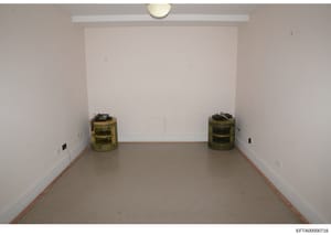 This is an evidence photograph showing an empty interior room with white walls and a concrete floor. Two cylindrical storage containers with olive/tan colored mesh material are positioned on opposite sides of the room. The room appears to be under re
