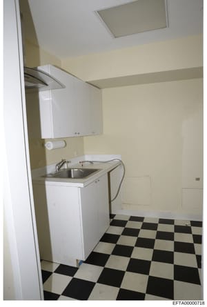 This is a photograph of a bathroom or utility room interior, showing basic fixtures and finishes. The image depicts cream-colored walls, white cabinetry, a stainless steel sink, and a distinctive black-and-white checkered tile floor. The photograph a