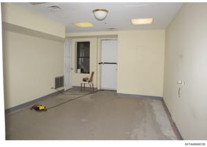 This is an evidence photograph showing an interior room in various stages of renovation or construction. The room appears to be part of a residential or commercial property, with bare concrete flooring, cream-colored walls, recessed lighting, and var