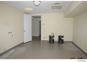 This is a photograph of an empty interior room, likely from an Epstein property documented during the investigation. The space shows a concrete floor, cream-colored walls, ceiling fixtures, and two black cylindrical objects in the center of the room.