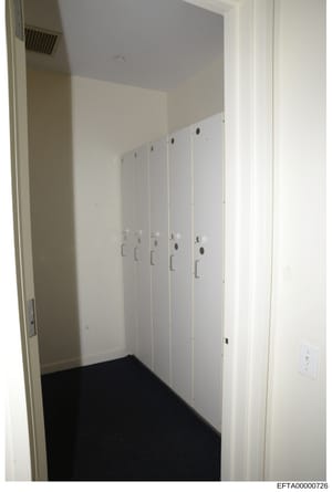 This photograph depicts the interior of a closet or storage room featuring five white locker-style cabinets with metal handles, mounted on walls with beige and blue paint. The room has black carpeting and basic electrical outlets, suggesting this is 