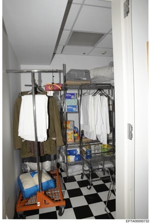 This is a photograph of an interior storage space containing household supplies, linens, clothing, and various packaged goods. The room features metal shelving units, an orange utility cart, white towels, clothing items, and boxes of household produc