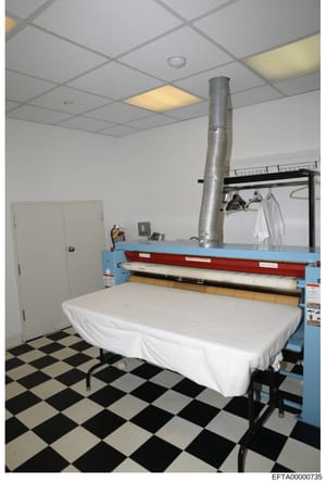 This is a photograph of a laundry or utility room, showing commercial-grade pressing/ironing equipment with a blue frame, white surface covering on a table, and a large metal ductwork pipe extending from the ceiling. The room has a checkered black an