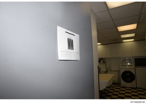 This is an evidence photograph showing the interior of a laundry room, likely from one of Epstein's properties. A document or chart is posted on the gray wall, and laundry appliances (washer/dryer) are visible on the right side. The image serves as v