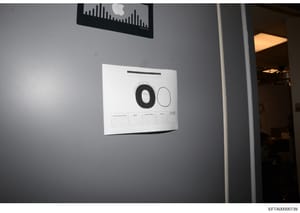 This is an evidence photograph showing a document posted on a gray wall, photographed with scale reference markers (numbered circles '0' and '00'). The document appears to be a form or record with a table below the scale markers. An Apple device is v