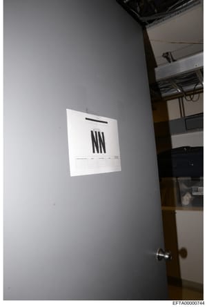 This is a photograph showing a white document or form posted on a gray interior wall, likely taken during an evidence collection or property search. The document appears to display 'NN' prominently and has a header and table structure typical of an o