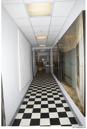 This is a photograph of a narrow, institutional-style hallway featuring a black and white checkered tile floor, white walls, drop-ceiling with recessed lighting, and what appears to be storage or utility items along the right side. The hallway appear