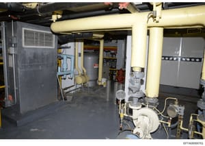 This is a photograph of a basement mechanical room, likely from an Epstein property, showing industrial heating, ventilation, and air conditioning (HVAC) equipment, piping systems, and utility infrastructure. The image documents the building's mechan