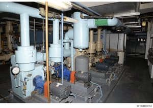 This photograph documents a building's mechanical room containing HVAC (heating, ventilation, and air conditioning) and plumbing infrastructure. The image shows industrial-grade equipment including white cylindrical tanks, blue and black pumps with o