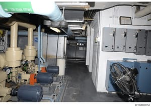 This photograph documents an interior mechanical/utility room containing HVAC equipment, including large tan-colored compressors or condenser units on the left side, multiple electrical control panels mounted on the right wall, ductwork, piping infra