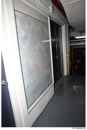 This is a photograph of a framed nautical or aeronautical chart displayed on a wall inside what appears to be an aircraft or vessel. The map shows coastal geography with blue water and gray landmass detail. The setting includes industrial or utility 