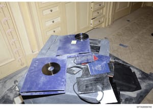 This is an evidence photograph showing items seized during a search, displayed on a table in what appears to be a residential interior. The image shows multiple blue passports, CDs/DVDs, electronic devices, and what appears to be crocodile or alligat