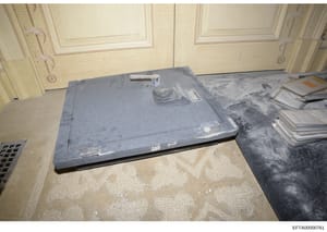 This is an evidence photograph showing a large gray metal safe or secure storage box on the floor of what appears to be an interior room with ornate ceiling details and cream-colored doors. The safe has visible locks and storage compartments on its s