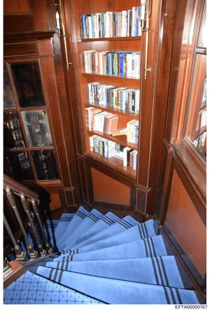 This is a photograph of an interior stairwell or hallway featuring a tall wooden bookcase with glass-fronted doors containing DVDs, CDs, and books. The image shows a blue striped staircase carpet and rich wood paneling, suggesting a high-end resident