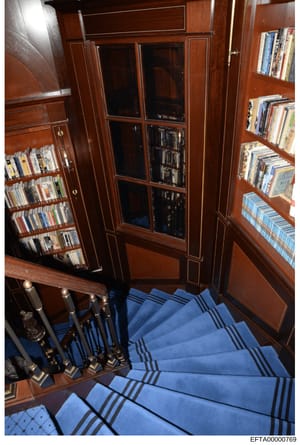 This photograph shows an interior view of what appears to be a luxury residential library or study space, featuring floor-to-ceiling wooden cabinetry with built-in bookshelves, glass-fronted display cases, and media storage. The room contains extensi