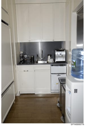 This is a photograph of a modern kitchen interior featuring white cabinetry, stainless steel appliances, and a dark countertop. The image appears to be evidence documentation from a property associated with the investigation, showing the condition an