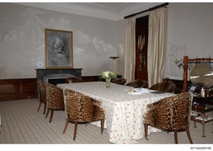 This photograph depicts an elegant formal dining room featuring a round dining table with a white floral tablecloth, surrounded by multiple leopard print upholstered chairs. The room contains period furnishings including a fireplace mantel with artwo